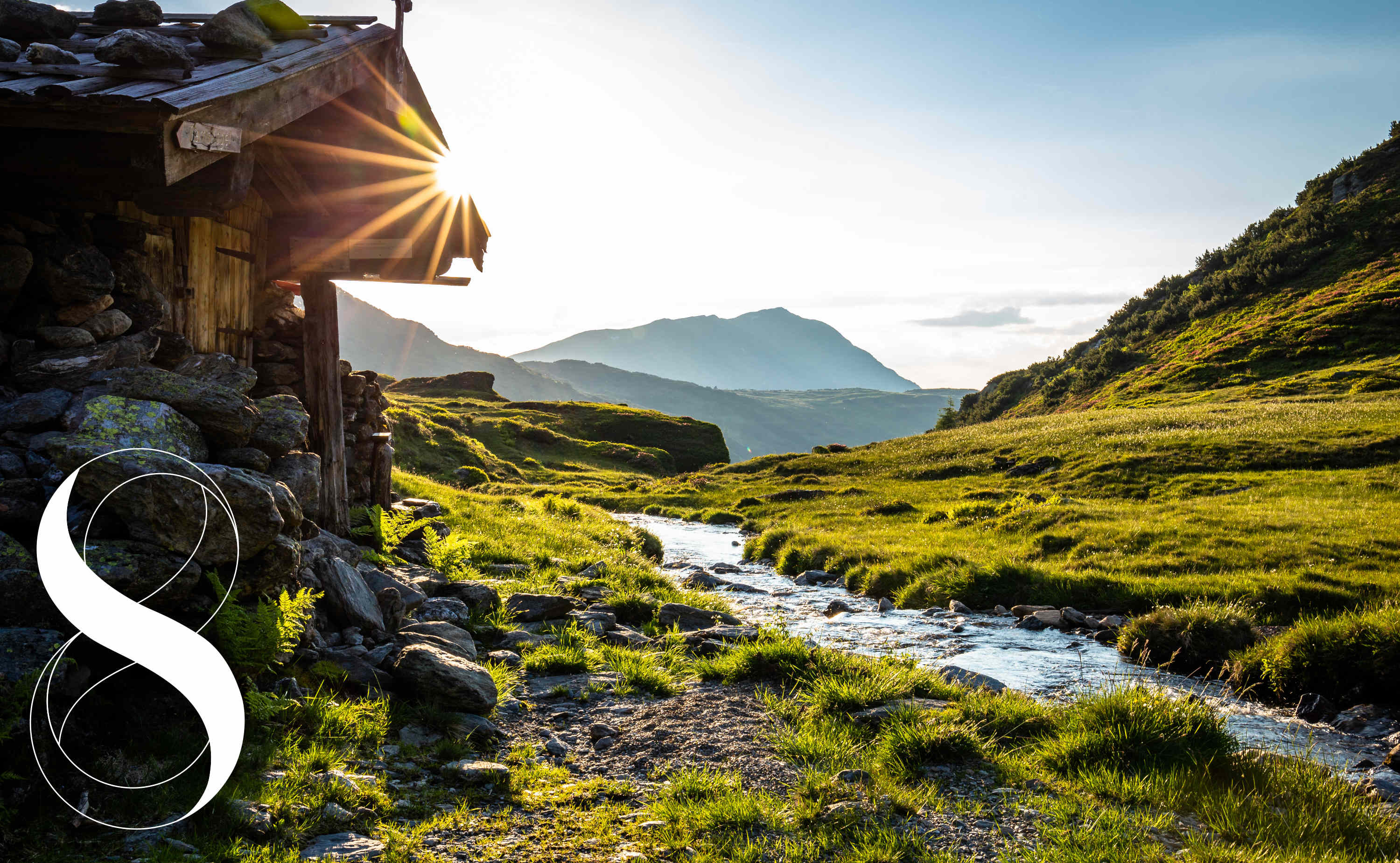 Eine kleine Hütte aus Holz und Stein steht neben einem klaren Bach. Die Sonne strahlt über die grünen Wiesen und wirft Licht auf das Tal. Im Hintergrund erheben sich Berge unter blauem Himmel – ein Symbolfoto für die Licht-Quell-Alm von Dr. Ewald Töth.