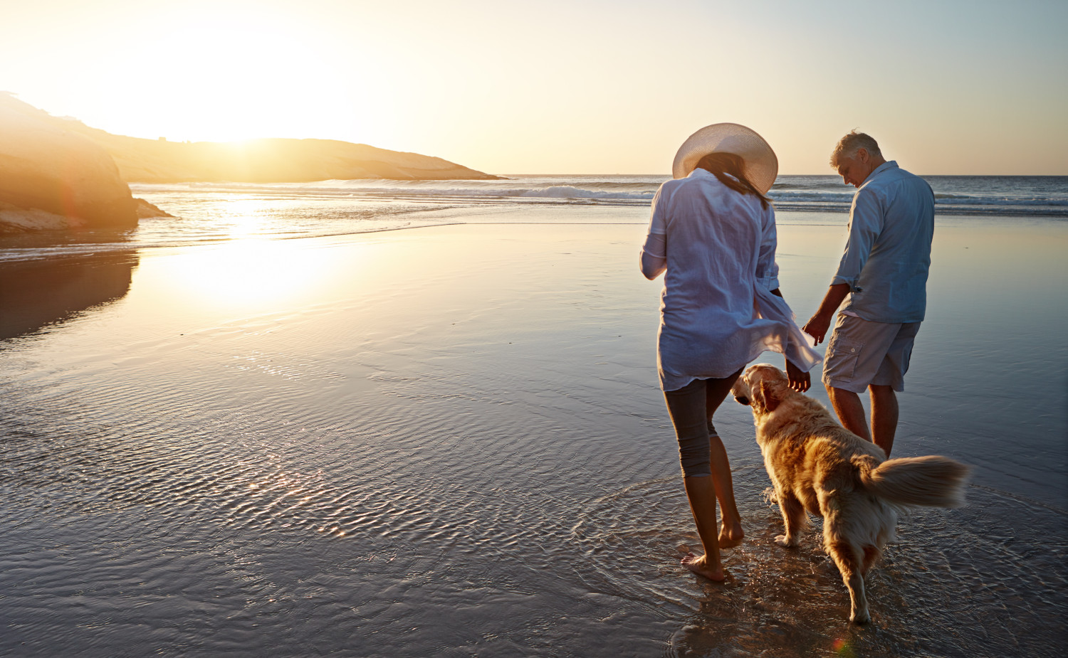 Ein Paar geht bei Sonnenuntergang mit seinem Golden Retriever am Strand entlang, die Sonne spiegelt sich auf dem nassen Sand und dem Wasser.