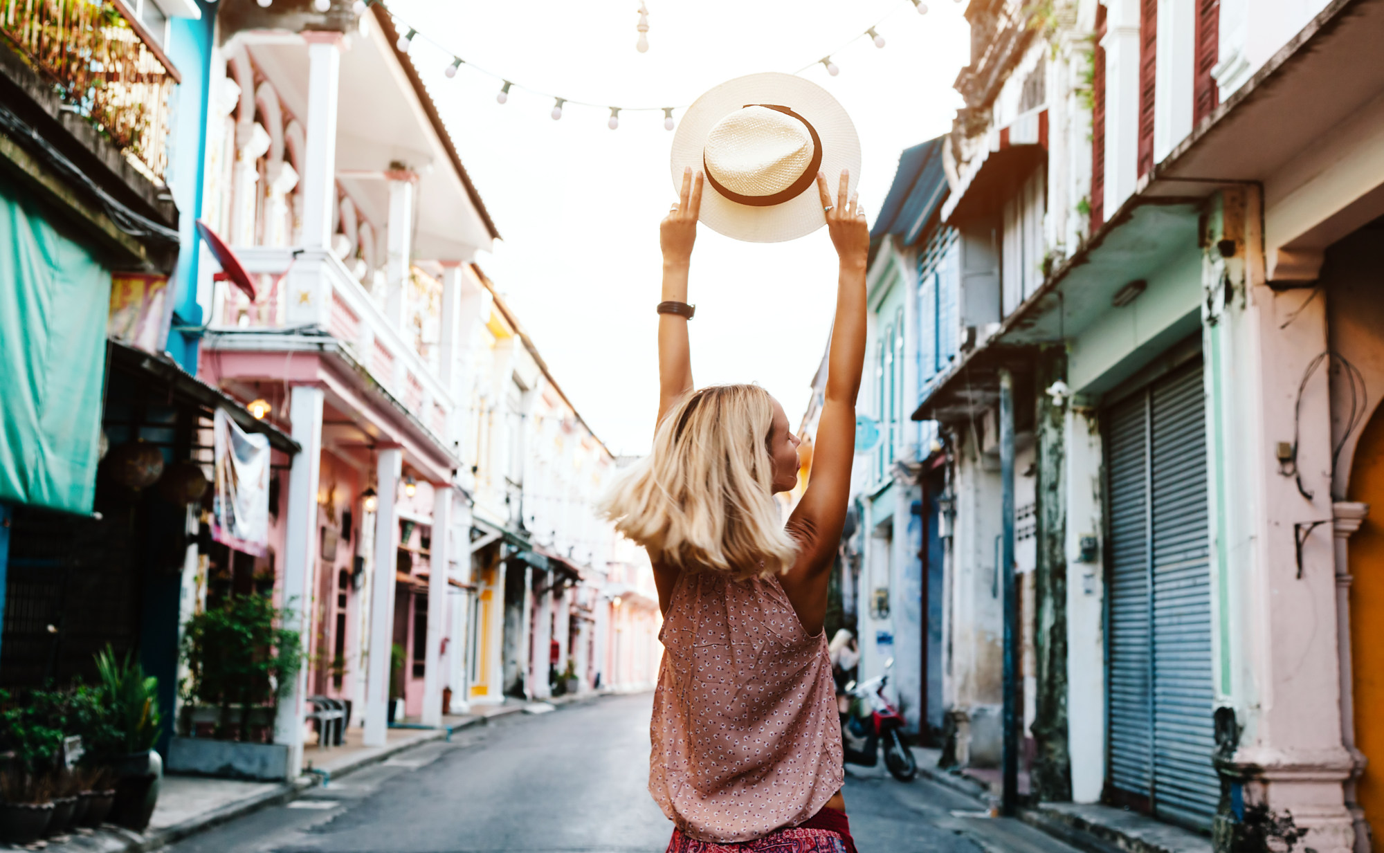 Eine Frau mit Strohhut, die ihre Arme auf einer Straße mit bunten Gebäuden in die Luft streckt. Ein Symbolfoto für ungebremsten Urlaubsgenuss ohne Krankheiten.