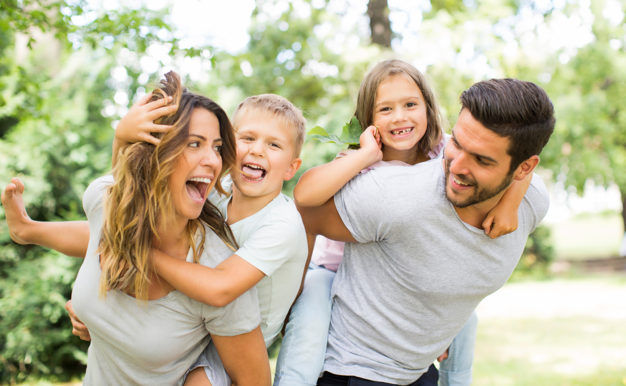 Eine lachende Familie im Freien, zwei Kinder auf den Rücken ihrer Eltern, Huckepack getragen. Alle vier scheinen Spaß zu haben. Symbolfoto für Freude, die auch durch Darmgesundheit entsteht.