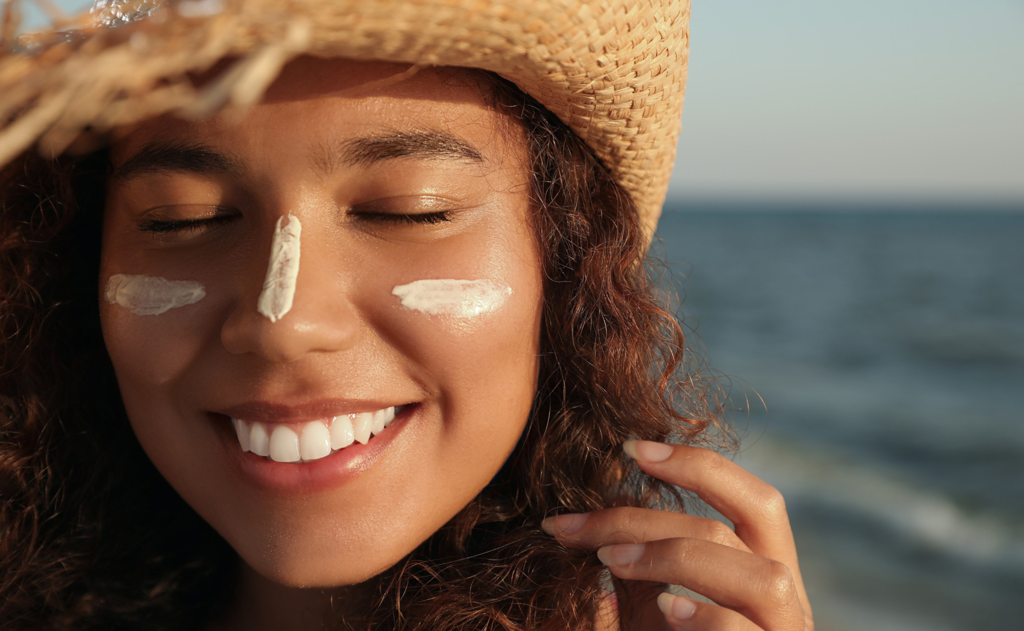 Eine lächelnde Frau mit braunen Locken und Strohhut hat Sonnencreme auf Nase und Wangen. Ihre Augen sind geschlossen, im Hintergrund ist das Meer. Symbolfoto für sonnengesunde Haut.